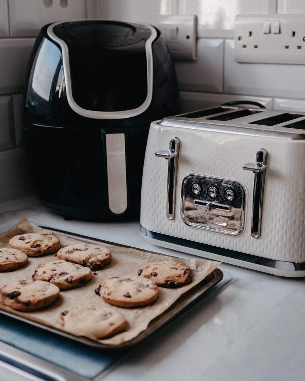 A black air fryer and a cream toaster on a kitchen counter, with a tray of cookies in front