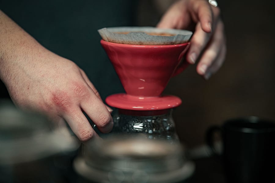 Hands setting a red ceramic pour-over cone and paper filter onto a glass carafe