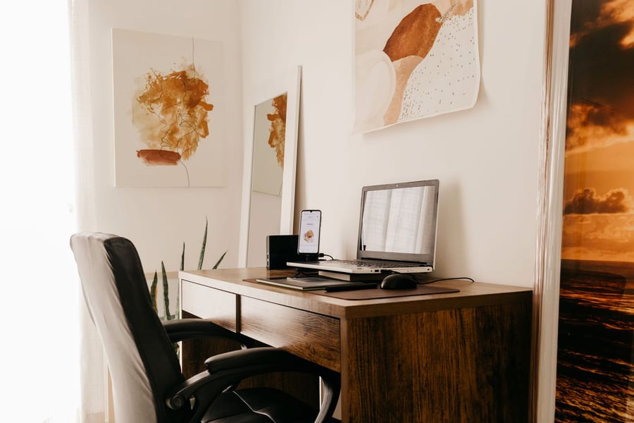 A wooden home-office desk with a laptop, a phone stand and an office chair beside a window