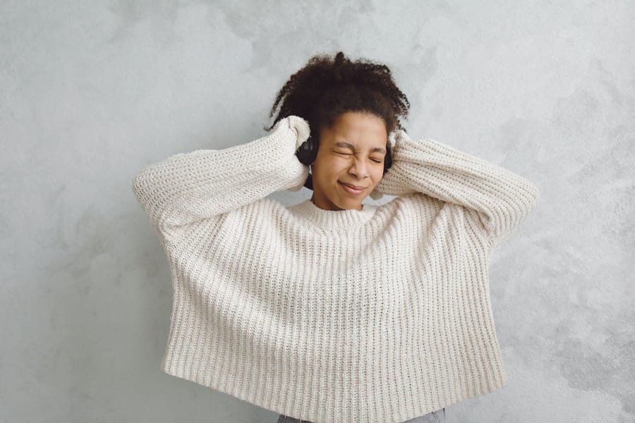 A woman in a cream knit sweater with her hands raised to the headphones she is wearing, against a grey wall